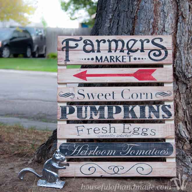 Decorative farmers market sign leaning up against a tree.