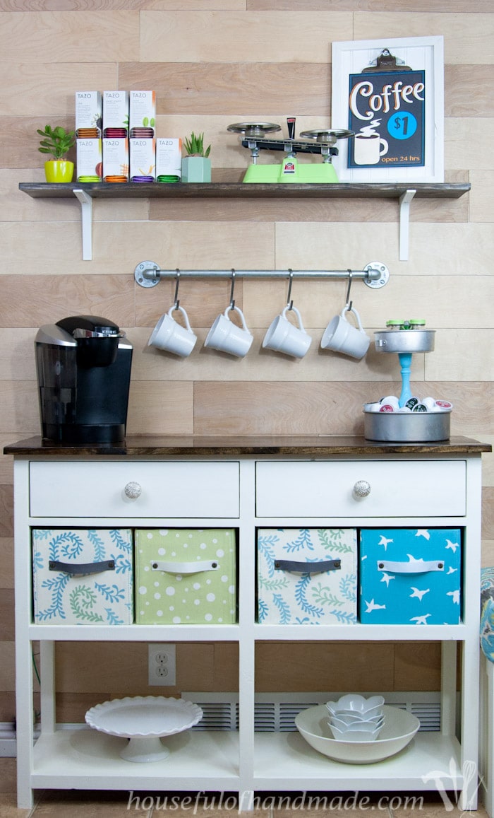 Coffee bar created on a white coffee station table with a shelf above it, hanging rack for mugs, fabric storage boxes, and k-cup storage. 