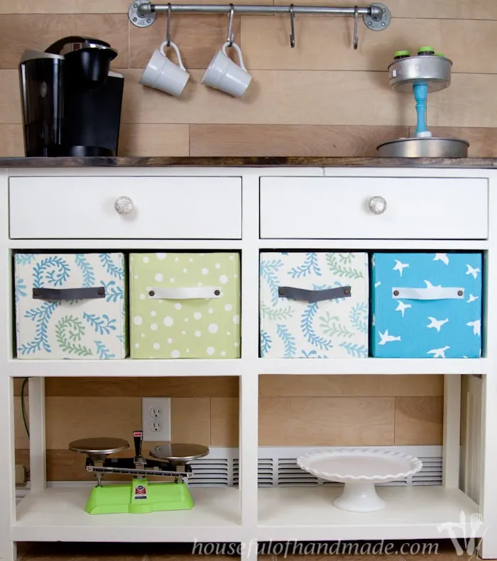 Fabric covered storage boxes on the shelf in a console table set up as a coffee center. 