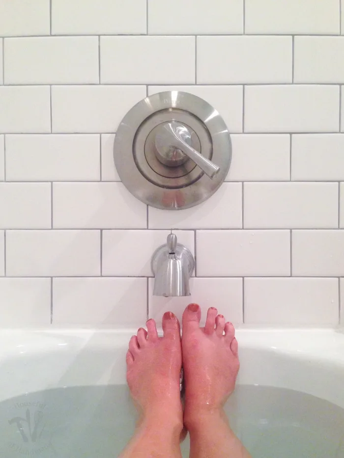 Feet sticking out of the bathtub in front of the faucet and subway tile surround. 