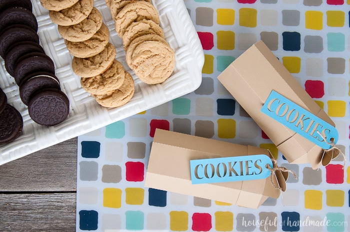 two printable paper cookie boxes laying horizontally on a table next to a tray of cookies