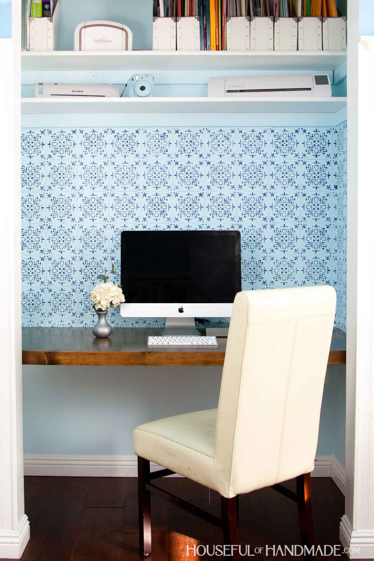 DIY desk in a closet with wood desk top, blue patterned wall paper with apple computer and desk chair.