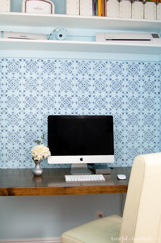 DIY desk built in a closet with wood desk top, blue wall paper for backsplash shown with white apple computer and desk chair.