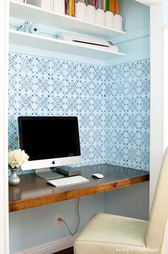 wood desk built in a closet with two shelves, desk chair and computer with blue patterned wall paper