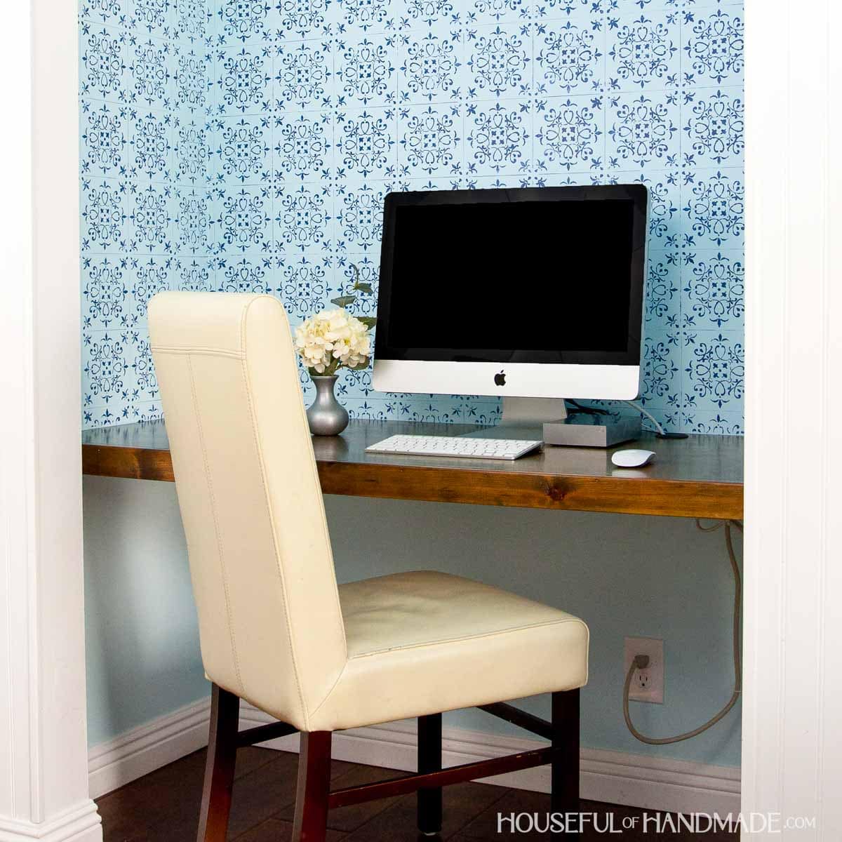 Office in closet shown with wood desk, office chair, blue wall paper and iMac computer.