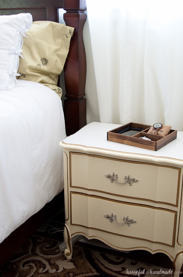 Vintage nightstand next to a bed with a wood tray holding a phone, earbuds, and watch on it. 