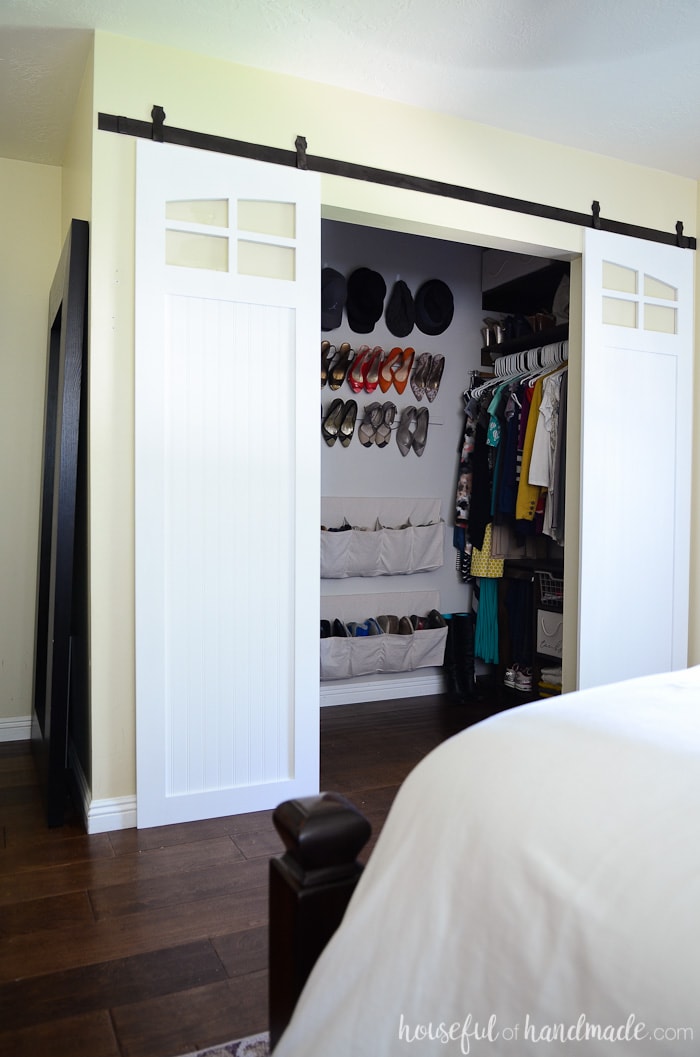 White closet sliding barn doors with window detail at the top in front of a closet with plywood storage organizers and hanging shoe storage. 