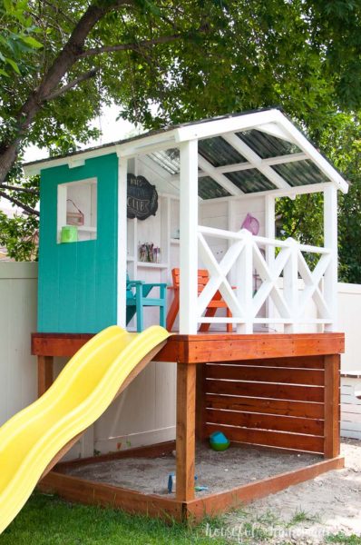 A small outdoor playhouse with X-railing, slide, walls and roof built over a sandbox.