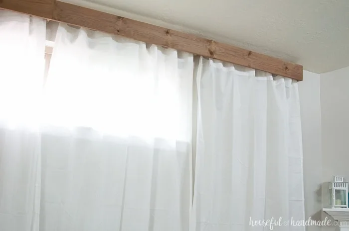 Finished window valance made from 1x6 boards installed on the wall with sheet white curtains hanging under it.