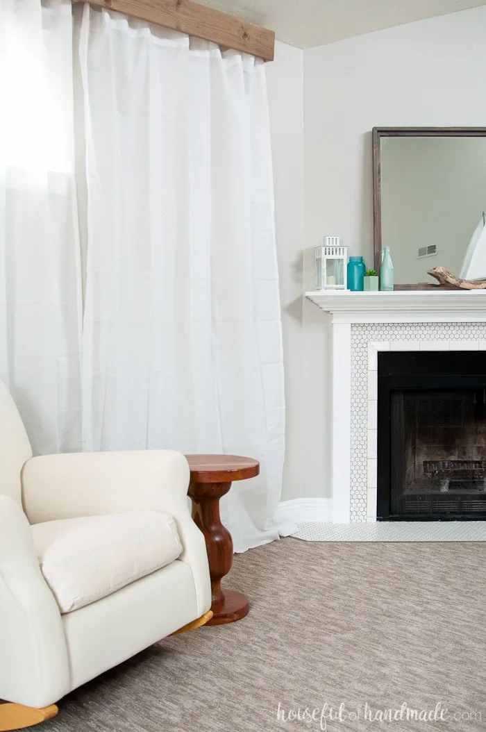 Living room with greige walls, a white hexagon tile fireplace and neutral rocker chair with a wood valance above the windows and white flowy curtains.