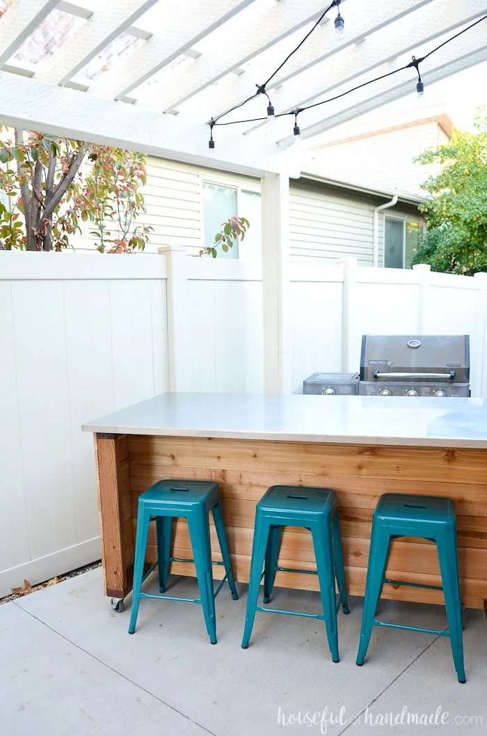 Cedar slat DIY outdoor kitchen island with on wheels next to a BBQ grill under a pergola. 