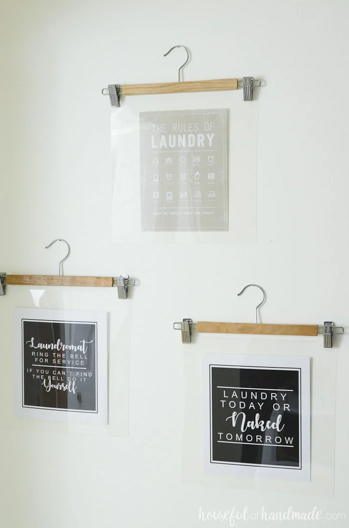 Printables hung on the wall with wooden hangers in a farmhouse laundry room. Housefulofhandmade.com