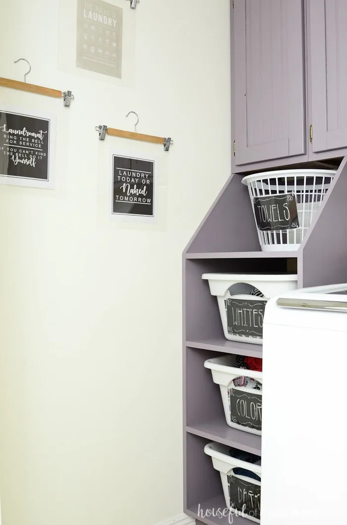 Purple stacking laundry basket storage with labeled laundry baskets. Art on the wall being hung with wooden pants hangers in a farmhouse laundry room. Housefulofhandmade.com