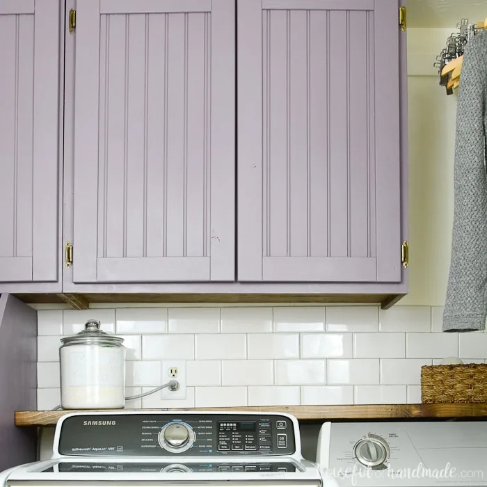 Purple painted beadboard cabinet doors with surface mounted hinges above a barnwood shelf behind a washer and dryer. 