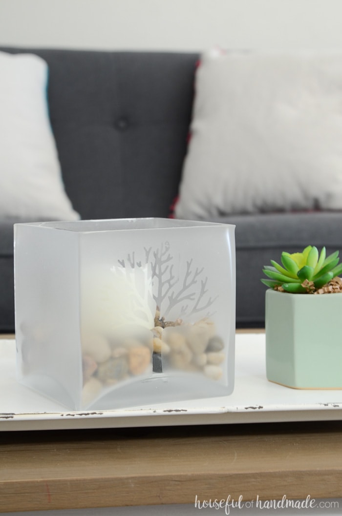 Decorate your home for spring with a large glass candle holder decorated with spring blossoms. This simple craft is the perfect way to shake the winter blues. Turn a boring glass candleholder into a beautiful piece of decor to transition the home into spring. Perfect for centerpieces or your spring mantel decor. Housefulofhandmade.com Frosted glass candle holder with tree design on a tray in front of a navy sofa. Easy DIY home decor. Housefulofhandmade.com
