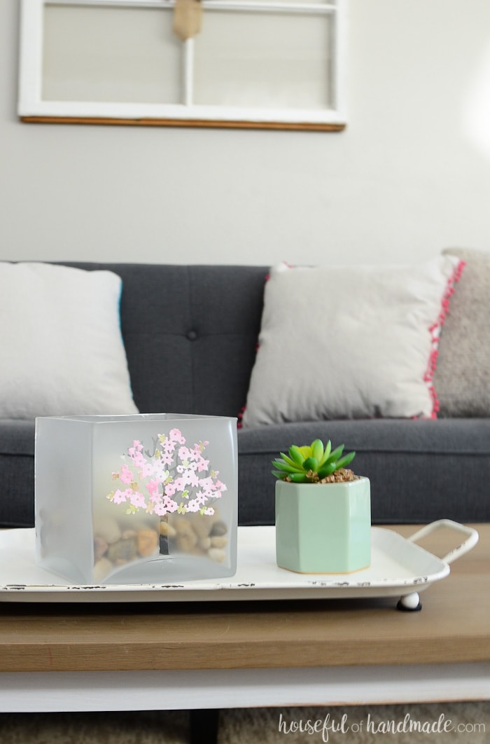 Coffee table with white tray and large glass candle holder with tree design. In front of a grayish-navy sofa with neutral pillows.