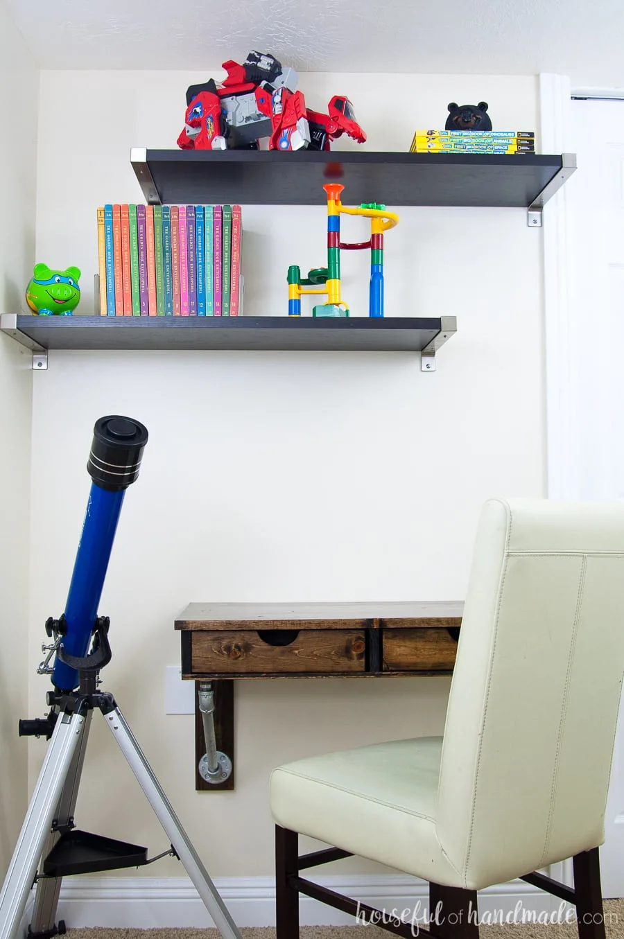 Wall mounted rustic desk in a boys bedroom. Shelves above hold encyclopedias and toys. Telescope sitting next to the desk.