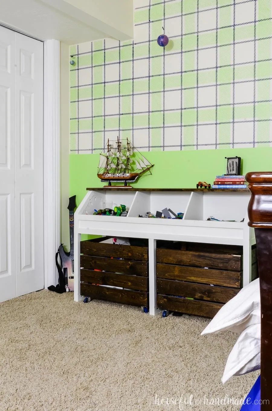 White and dark wood toy storage unit with open bins on the top and large rolling bins underneath. In a boys bedroom with a green plaid painted wall and neutral beige carpet.