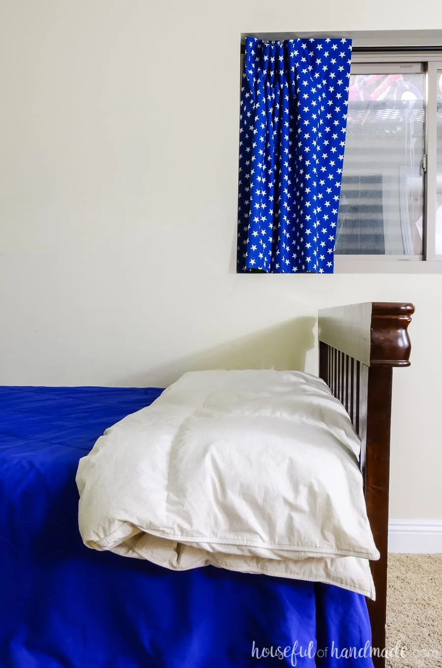 The foot of the bed with a dark wood craftsman footboard and white down comforter. Black out curtains in the window with blue fabric with white stars on it. 