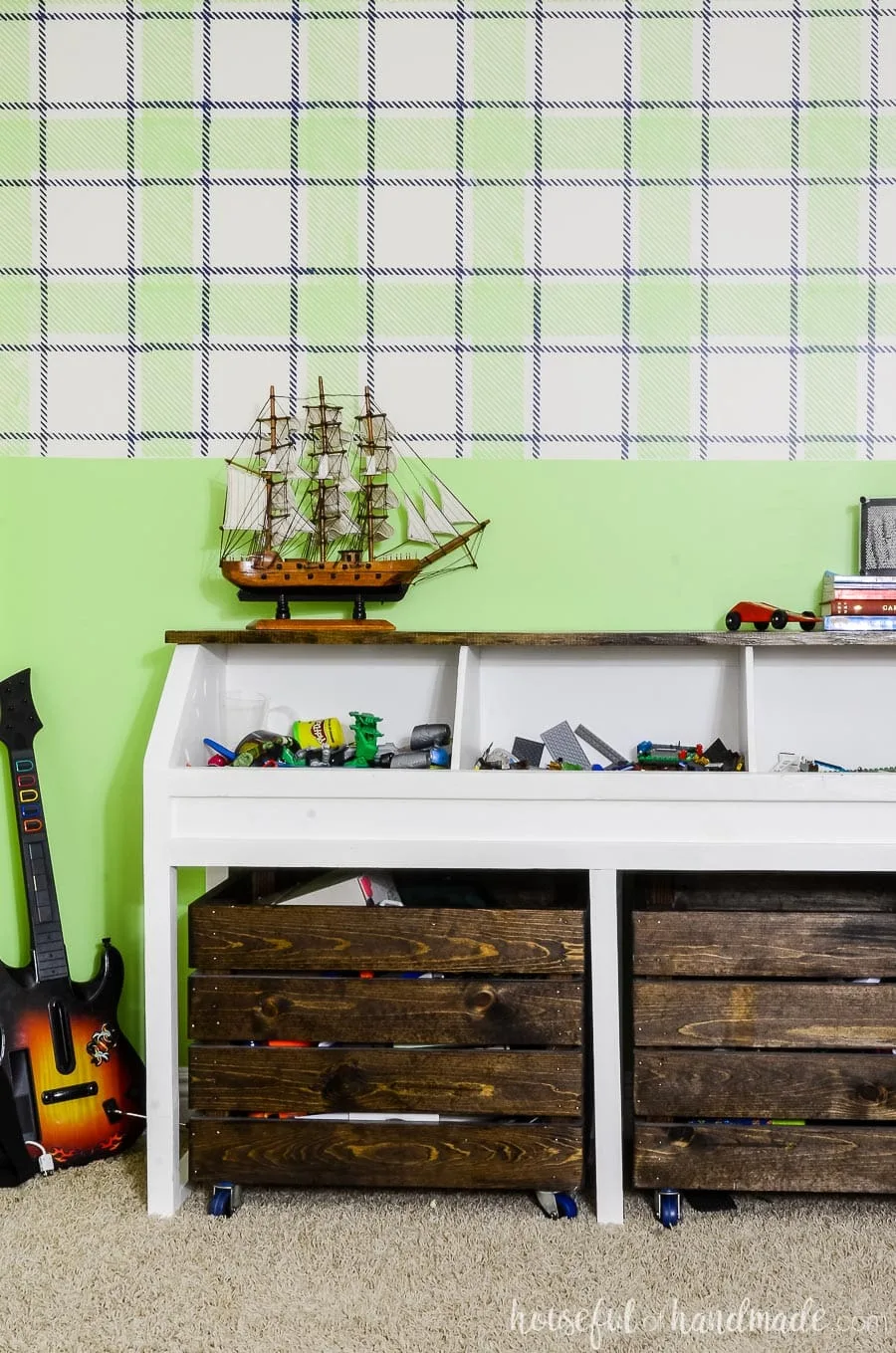 Rustic toy storage console full of Legos and action figures. In front of a statement wall painted with with a green & navy plaid design on top and green solid on the bottom. Wii guitar in the corner. 