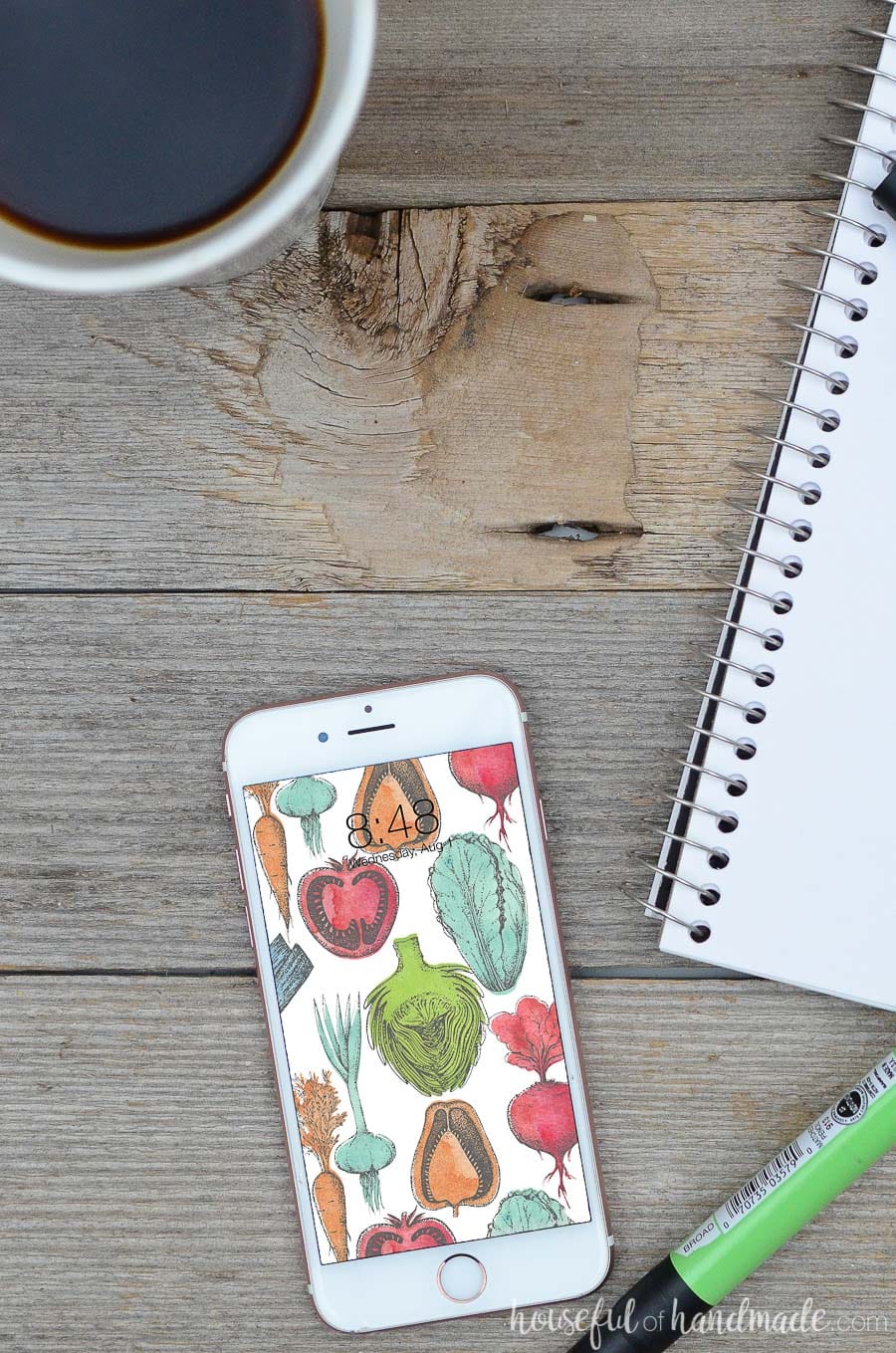 Bring the farmers market to your screens with these beautiful free digital backgrounds for August. The watercolor vegetable pattern is the perfect way to decorate your electronics for the last days of summer. #digitalbackground #digitalwallpaper #vegetableart #freedownload White iPhone smartphone with colorful vegetable design digital wallpaper on it. Phone is laying on a wood background with sketchbook and cup of coffee next to it.