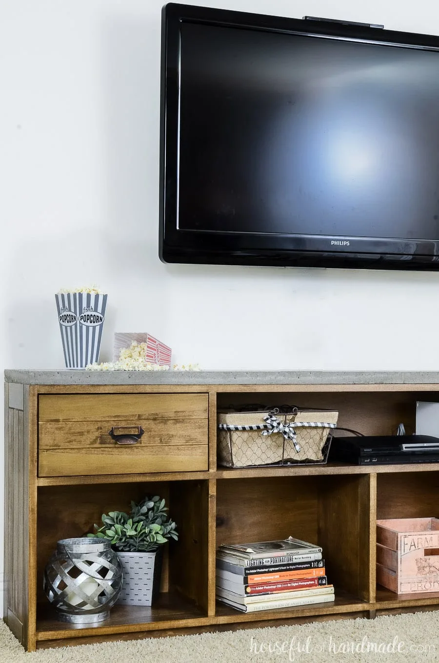 Televison mounted on the wall above a warm brown rustic TV stand with concrete top. Showing open shelving with books and decor items inside.