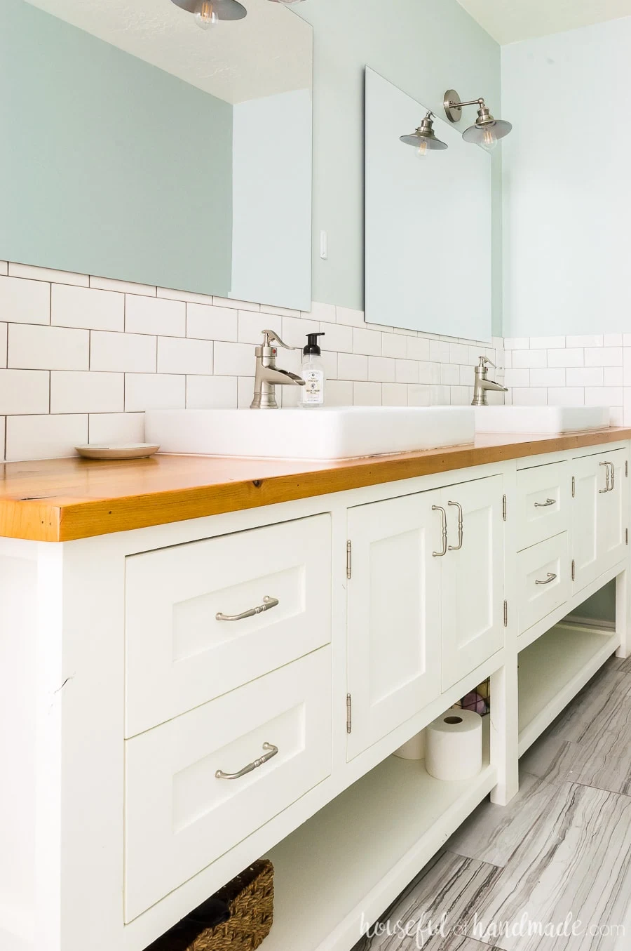 A DIY master bathroom retreat with a large 8' white mission style open shelf bathroom vanity with natural wood vanity top. White square vessel sinks and white subway tile on backsplash. Frameless mirrors against gray-green walls make a peaceful space.