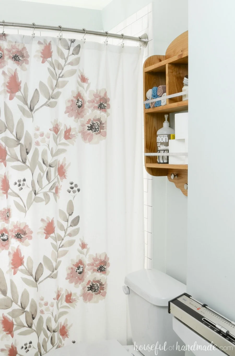 Wood over toilet shelves storing toilet paper and other bathroom items. Next to watercolor floral shower curtain. 