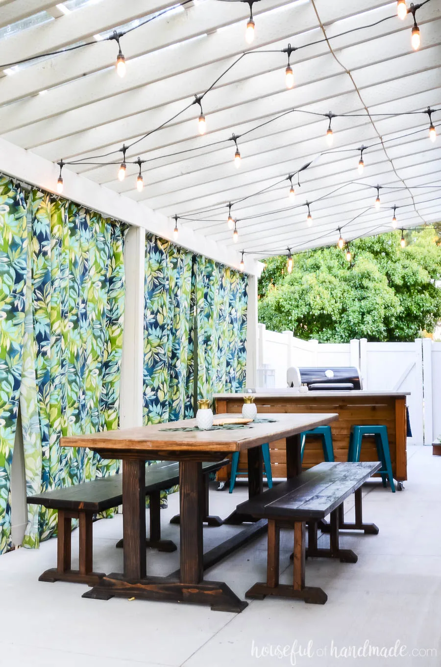 White pergola with lights over a large outdoor dining table with benches. Outdoor kitchen island next to the grill in the background.