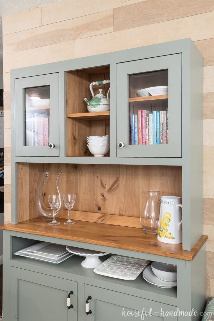 Natural wood hutch with painted gray interior storing platters, cook books, and baking dishes.