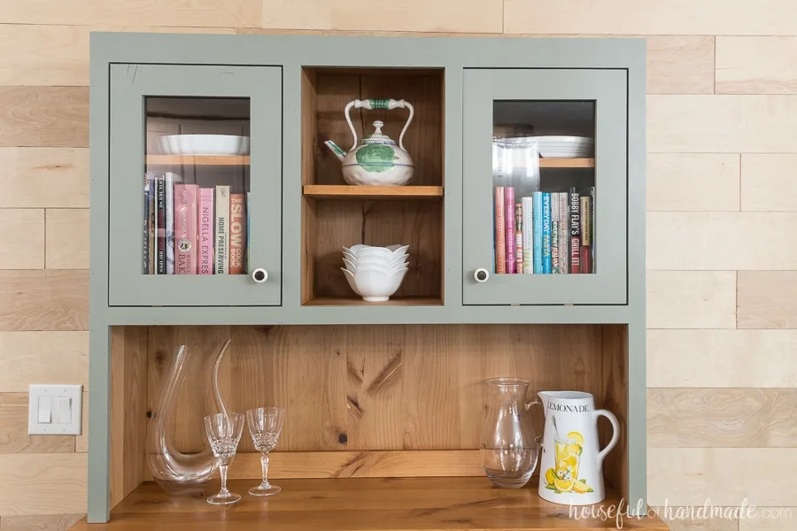 Natural wood hutch with painted gray interior storing platters, cook books, and baking dishes.