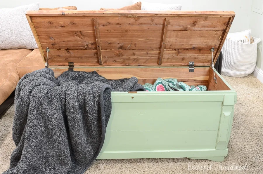 The refinished storage chest with the lid open showing all the blankets inside. One gray fluffy blanket draped out over the corner of the chest. 