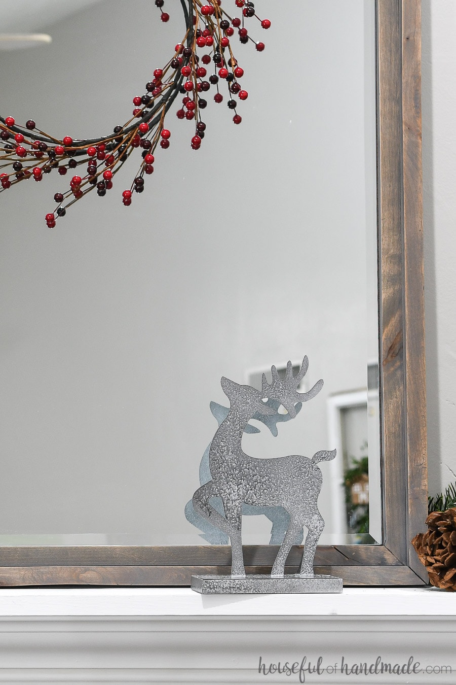 One paper reindeer decorations in front of a mirror under a red berry wreath.