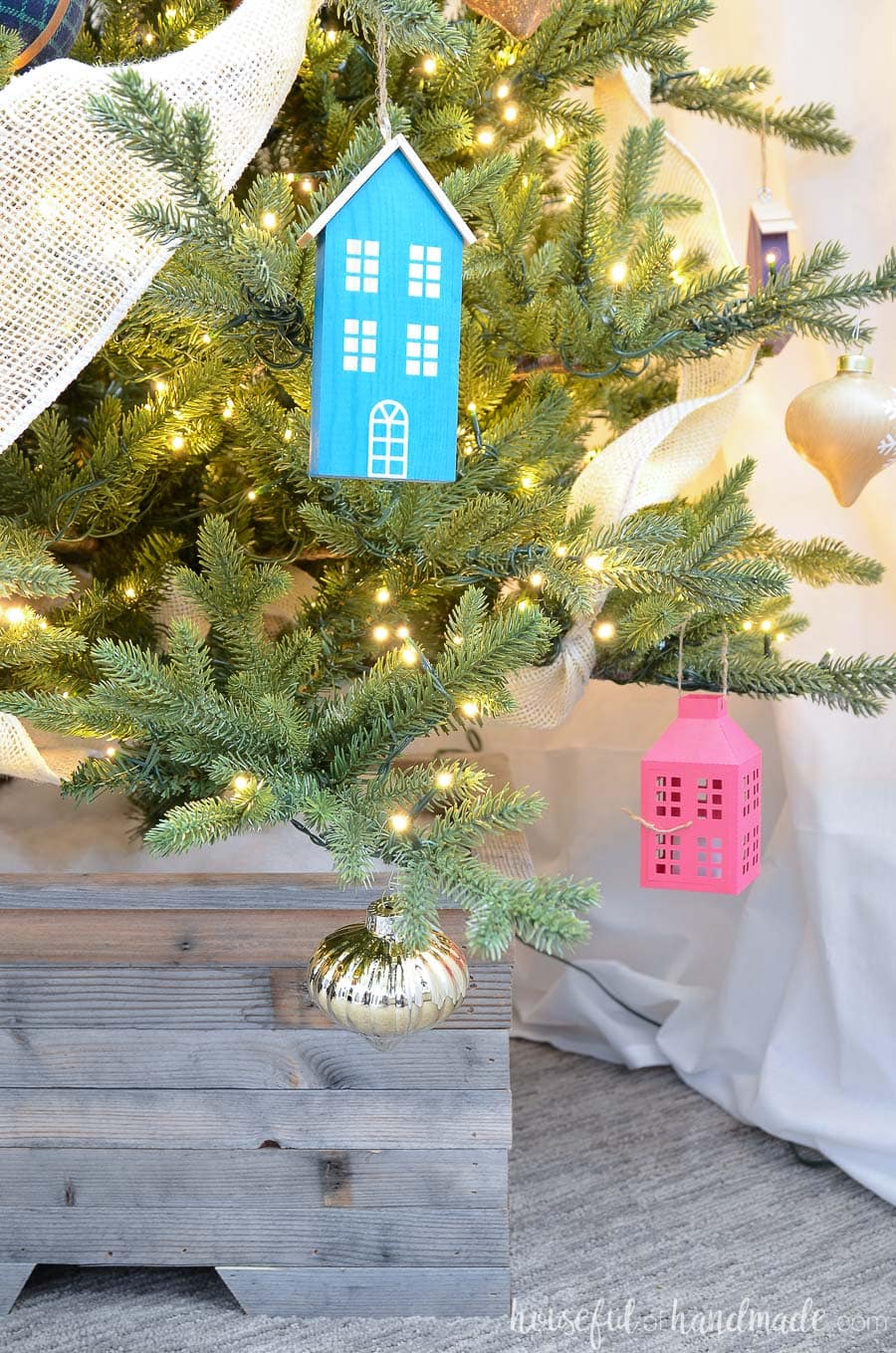 Wood house ornaments and paper lantern ornaments on a Christmas tree inside a wood tree collar. 