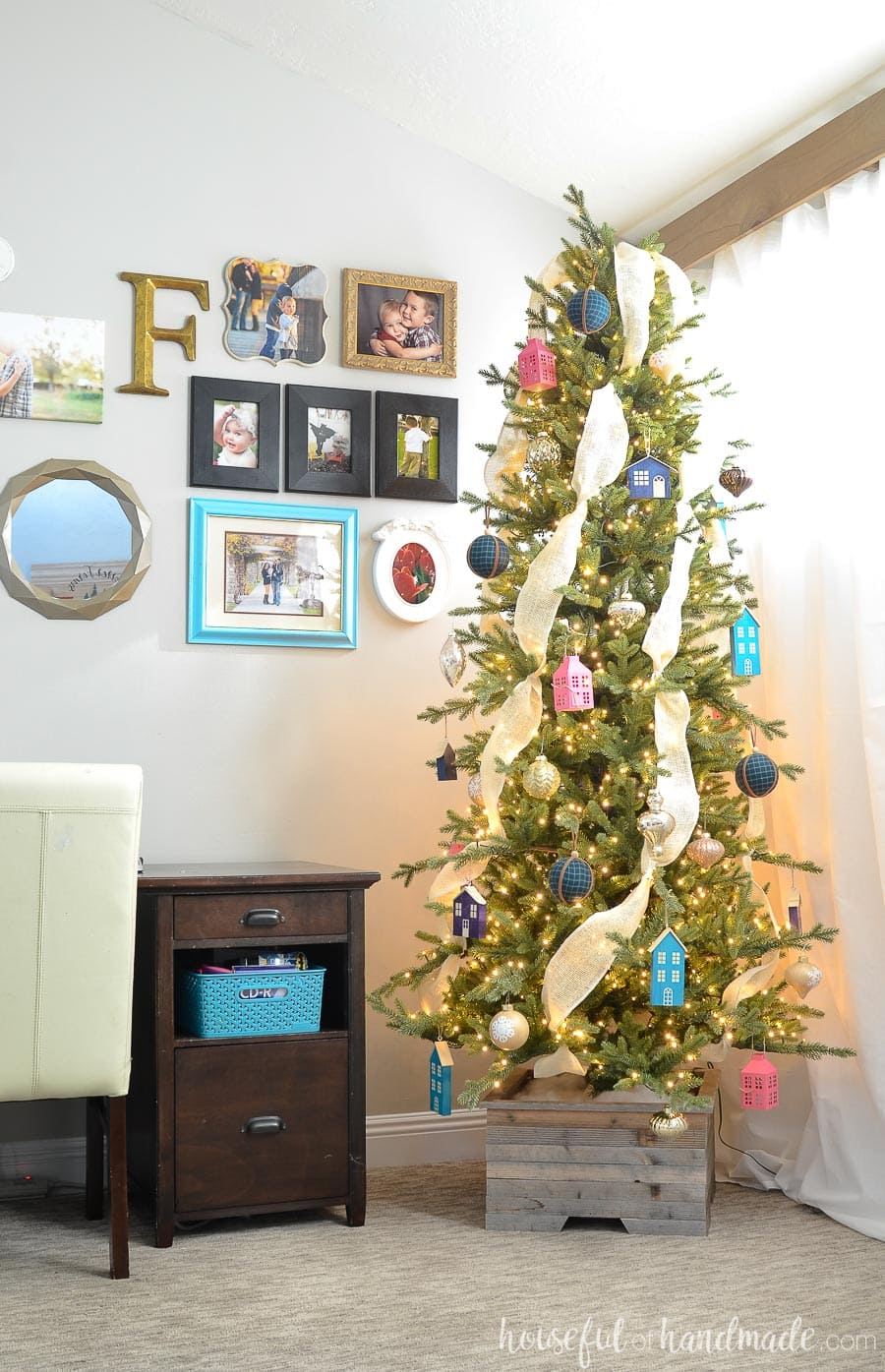 Christmas tree in a wood Christmas tree stand with bright colorful ornaments next to a desk.