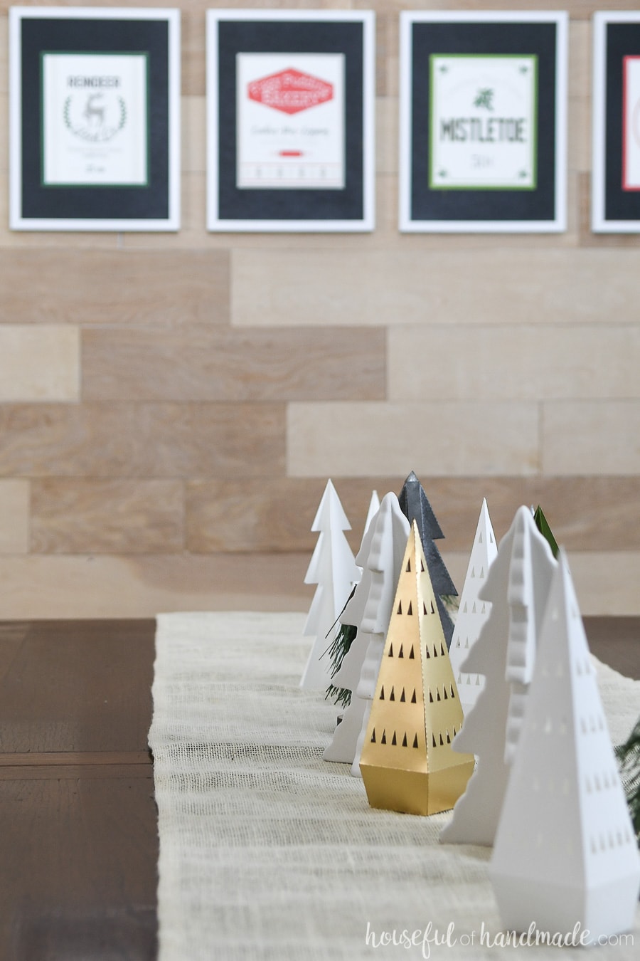 Dining room table filled with a variety of paper Christmas trees and framed printable Christmas signs in the background. 