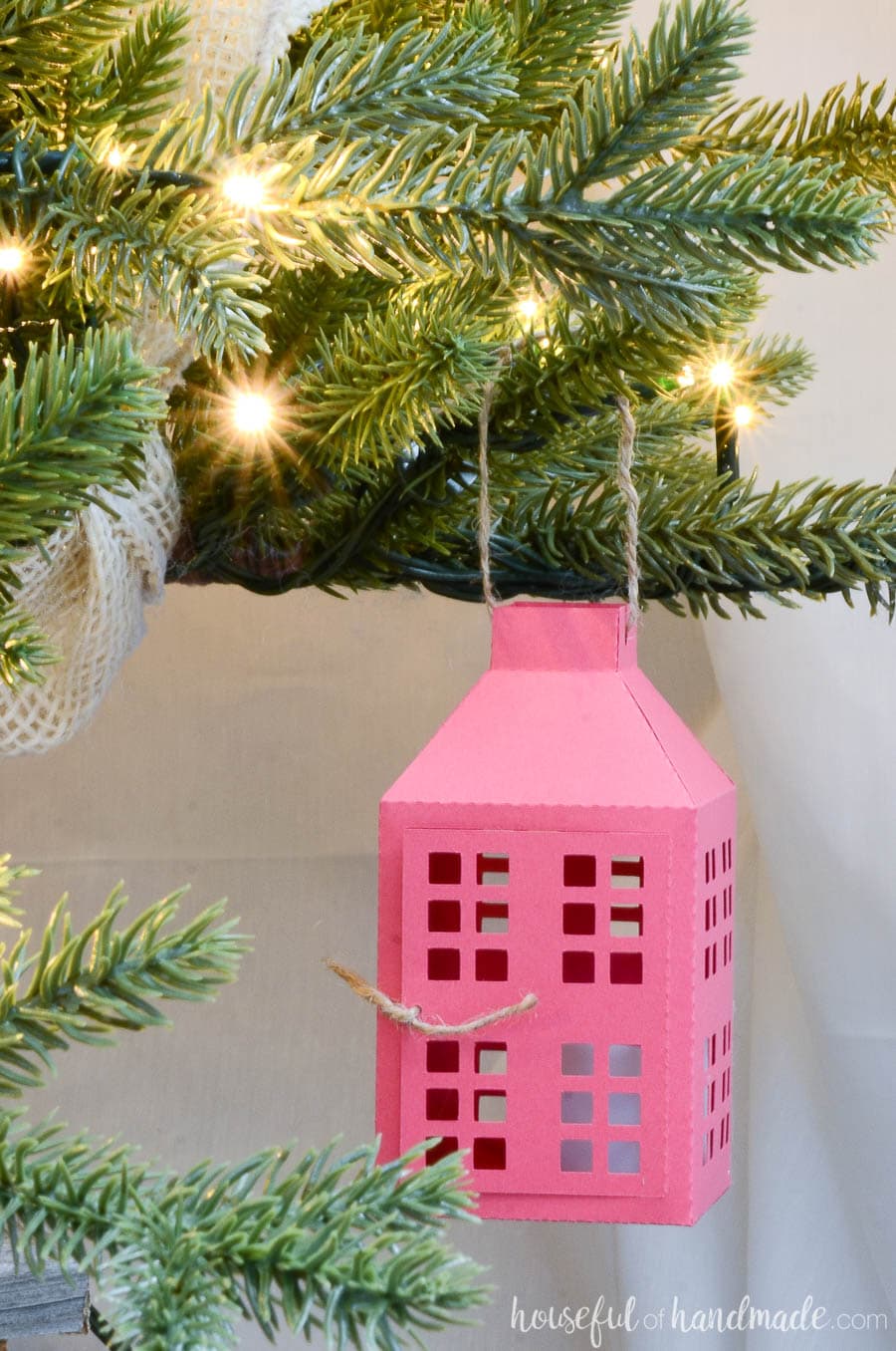 Paper Christmas tree ornament in the shape of a farmhouse lantern hanging on the branches of a Christmas tree. 