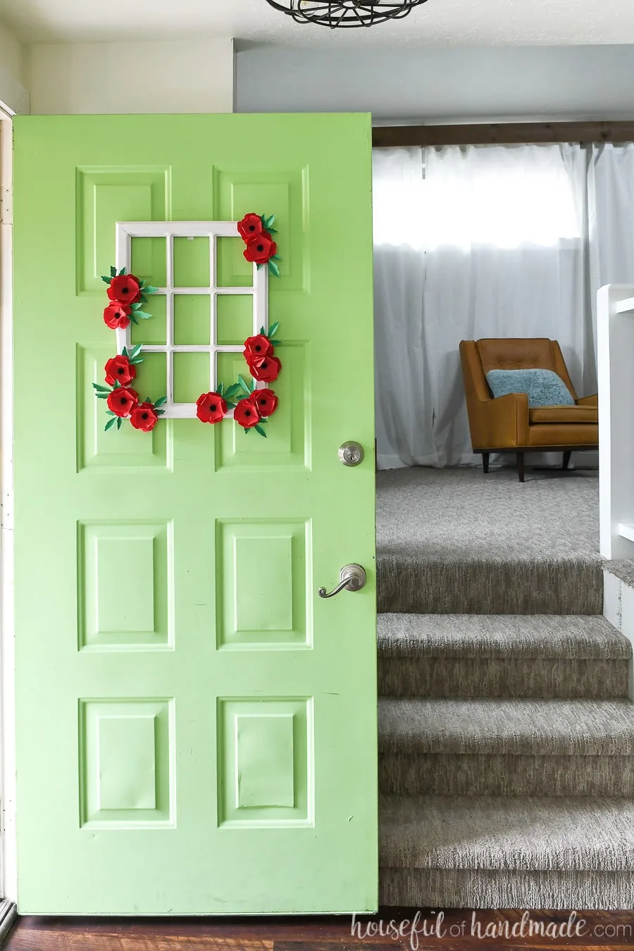 Green door with window frame wreath on it, decorated with paper poppy flowers, opening up to the house.