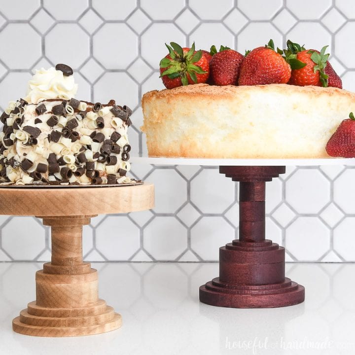Two DIY wooden cake stands with cake on them in front of a white tile backsplash.