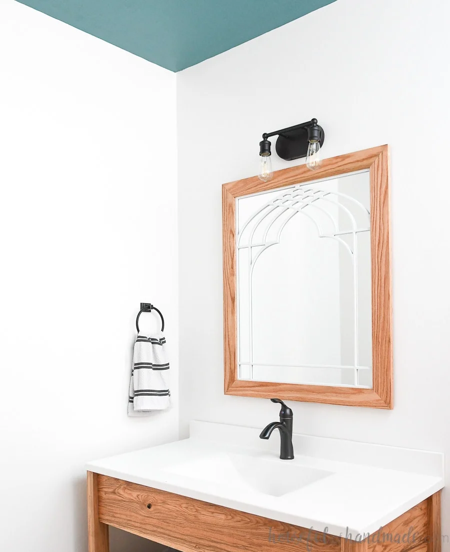 Bathroom with wood vanity, white vanity top, window frame mirror, antique bronze fixtures, and teal painted ceiling. 
