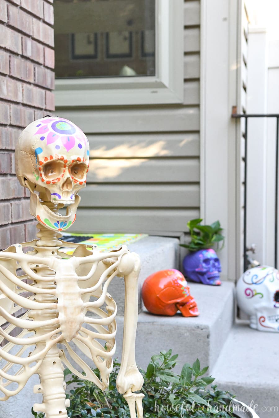 Halloween skeleton decorated to look like a sugar skull in front of porch decorated with more sugar skulls. 