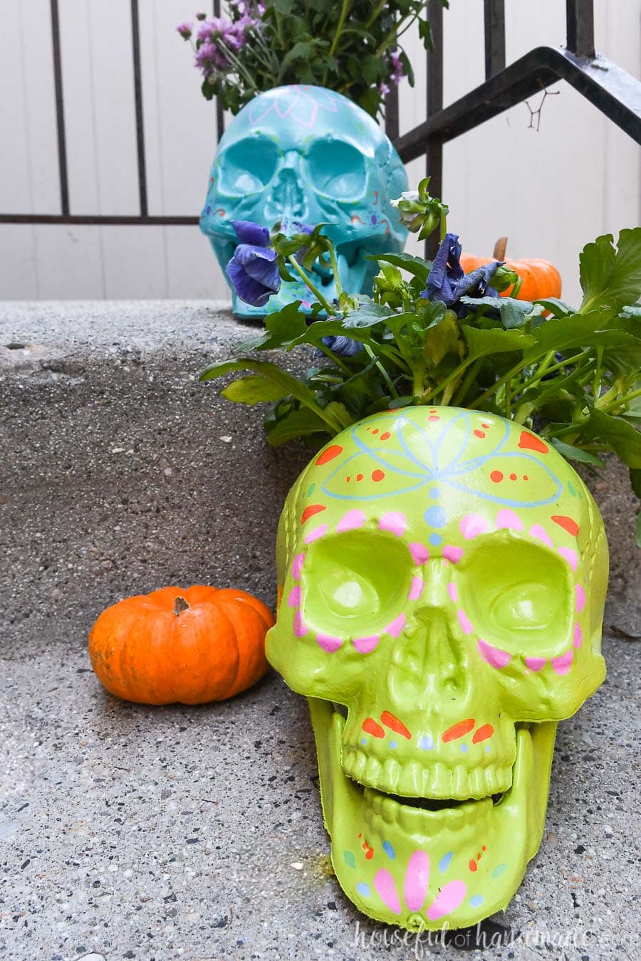 Green and blue sugar skull planters holding fall flowers on a porch. 