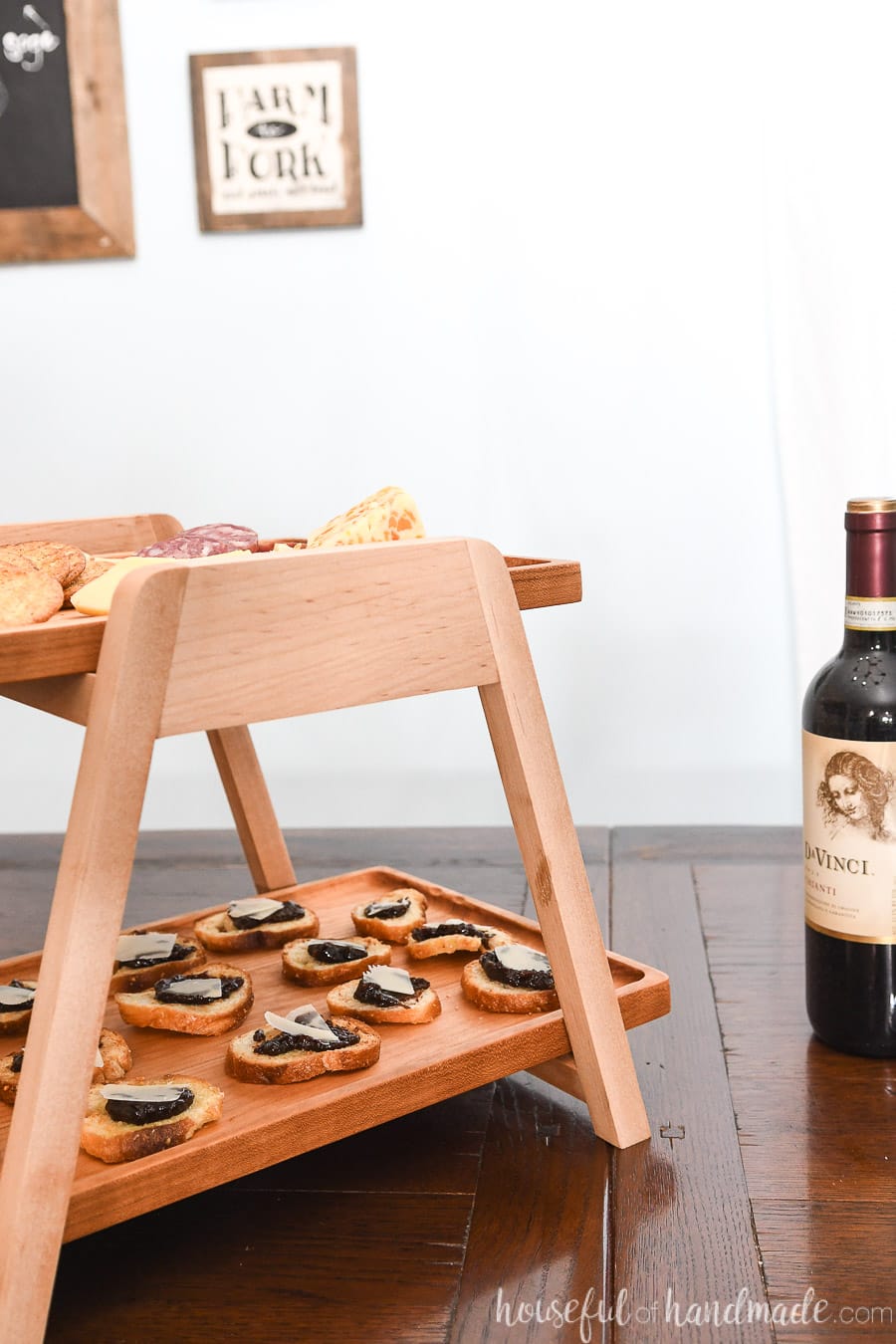 Two tier wood serving tray with appetizers on it next to a wine bottle. 
