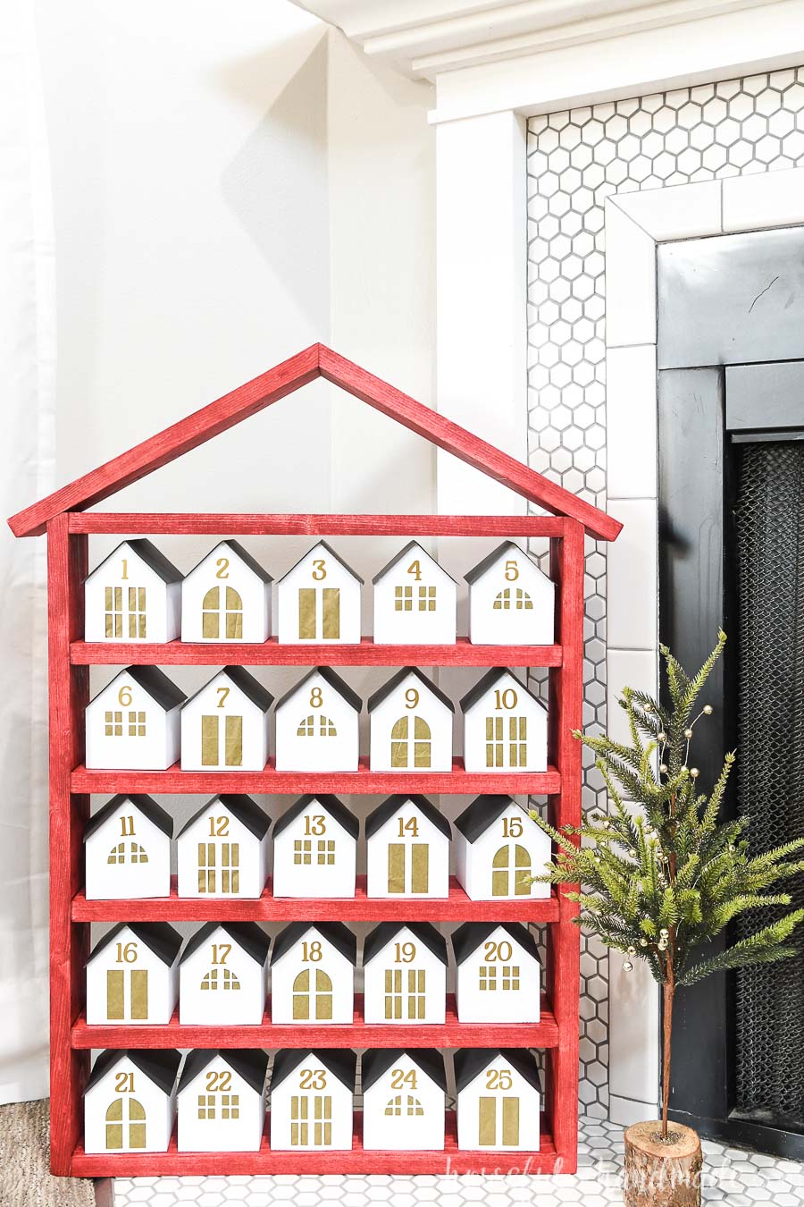 Red house shaped shelf with 25 white & gold paper houses on them in front of a fireplace.