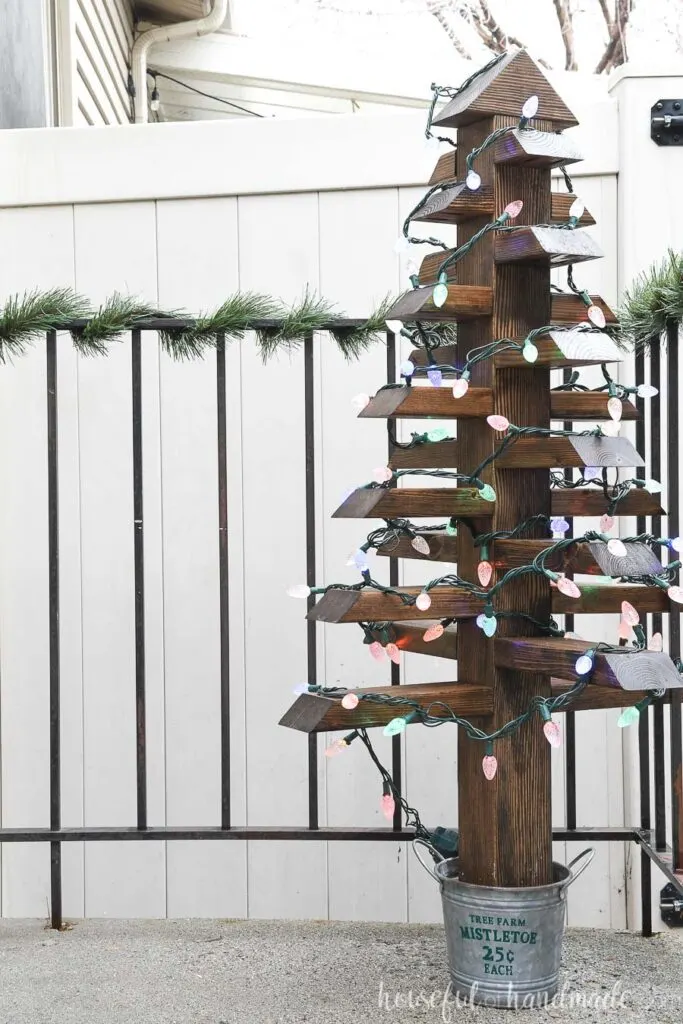 Outdoor Christmas tree with lights on the porch by a railing wrapped in garland.