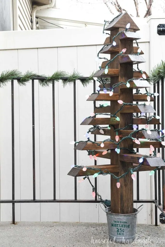 Outdoor Christmas tree with lights on the porch by a railing wrapped in garland. 