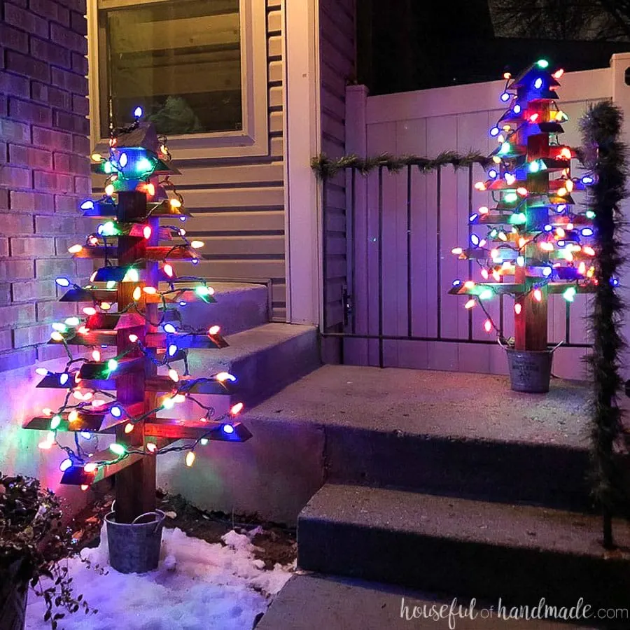 Two 2x4 Christmas trees with colored Christmas lights on the porch at night. 