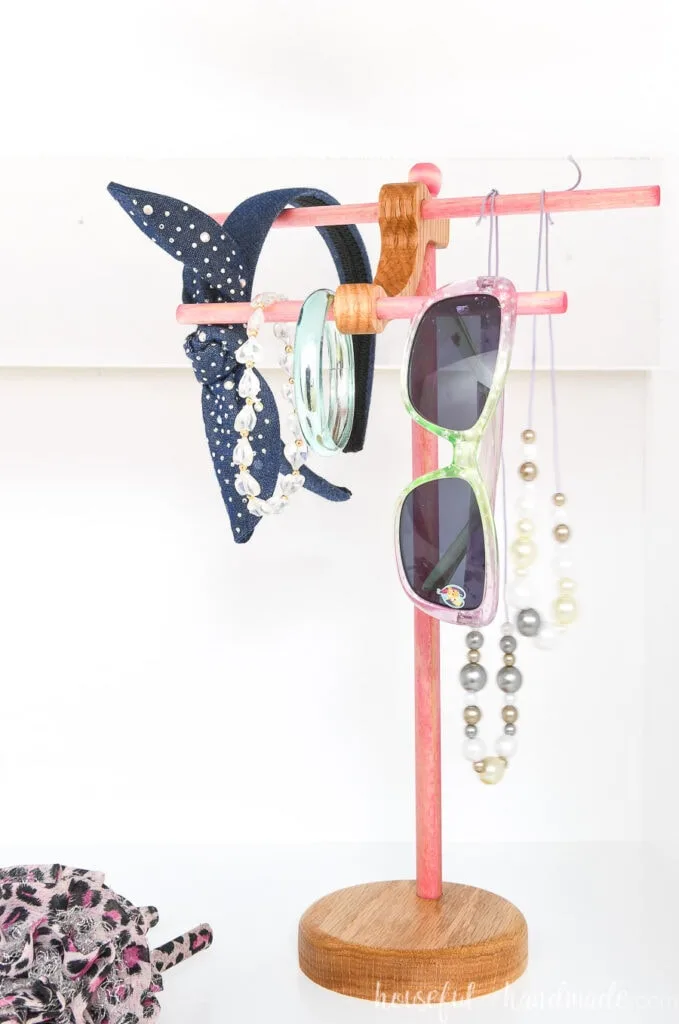 Pink and wood stained necklace holder with kids necklaces, sunglasses, and headbands on it. 