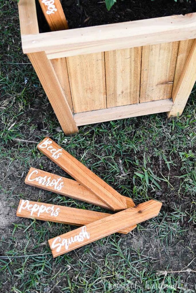 Four cedar garden markers with white acrylic inlays laying on the ground in front of a planter.