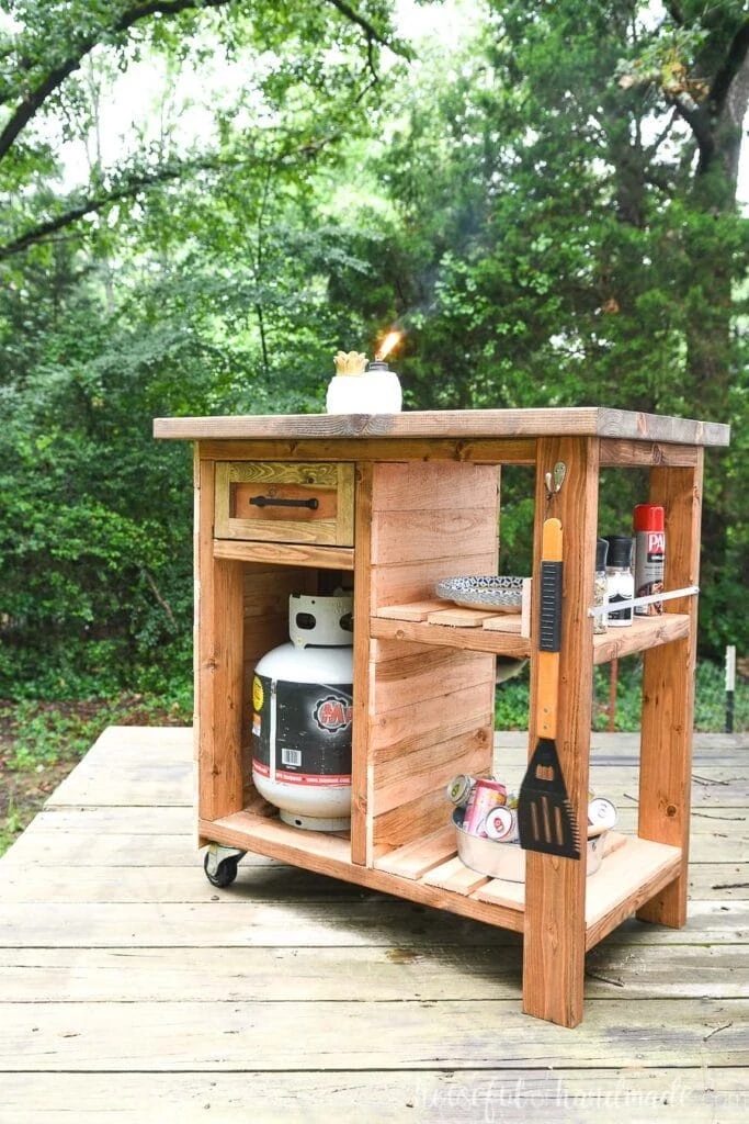 Cedar clad rolling grill cart with storage sitting on a deck in front of trees. 
